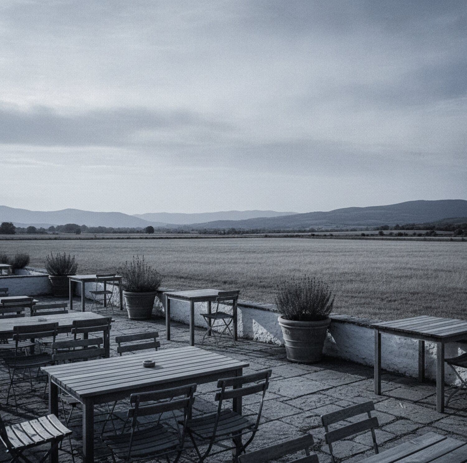Leere Terrasse mit einfachen Holztischen und Kräutertöpfen, dahinter offene Wiesen und die ferne Linie der Voralpen